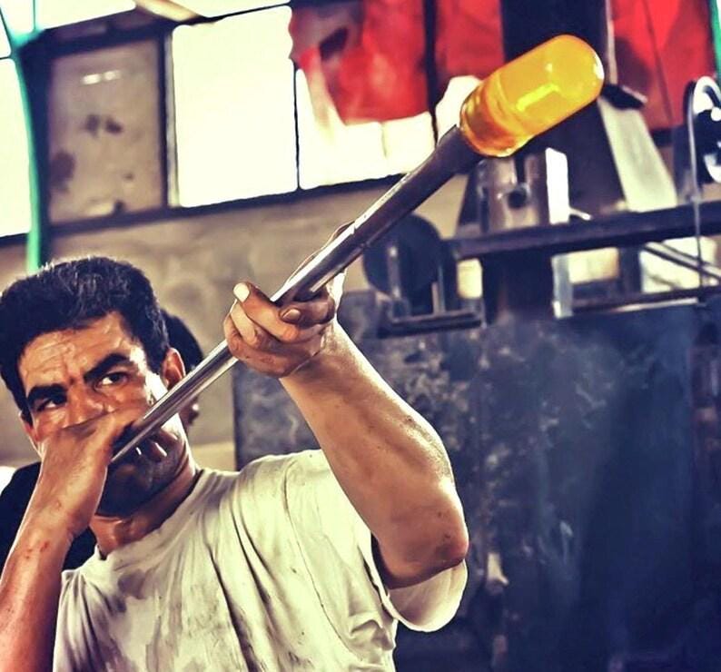 Artisan glassblower shaping molten yellow glass on a long metal rod in a workshop, House of Anouar